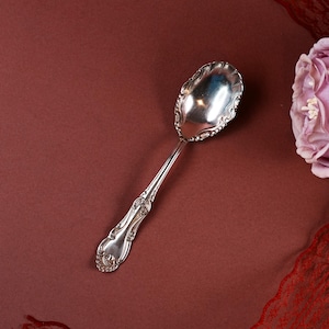 Op de afbeelding: Een zilveren lepel met een sierlijk handvat en een decoratieve, geschulpte kom. De lepel staat tegen een dieprode achtergrond, met een vleugje roze bloem en rood kant in de hoek.