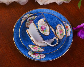 Könnte beinhalten: Ein blaues Vintage-Teeservice mit Goldrand, bestehend aus einer Teetasse, Untertasse und Kuchenteller. Das Set hat ein Blumenmuster mit rosa, gelben und grünen Details. Die Innenseite der Tasse ist weiß.