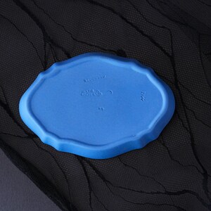 May include: A matte blue oval ceramic tray with a scalloped edge. The underside features embossed text reading WEDGWOOD, MADE IN ENGLAND, and the letters DW. The surface has a smooth, uniform finish with a distinct moulded rim and central markings.
