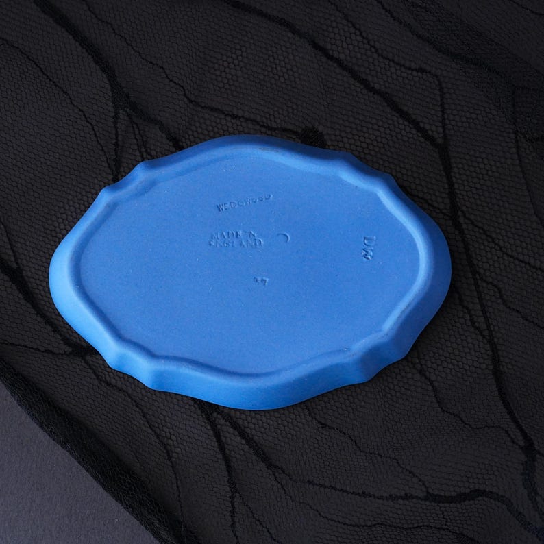 May include: A matte blue oval ceramic tray with a scalloped edge. The underside features embossed text reading WEDGWOOD, MADE IN ENGLAND, and the letters DW. The surface has a smooth, uniform finish with a distinct moulded rim and central markings.
