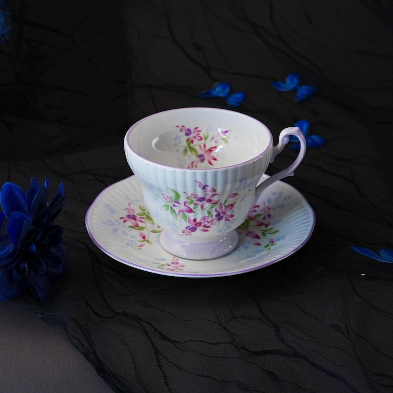 May include: A white porcelain teacup and saucer set featuring a ribbed texture and a delicate floral pattern of pink and purple fuchsia flowers with green leaves. The rim of both the cup and the saucer is accented with a thin, light purple trim.