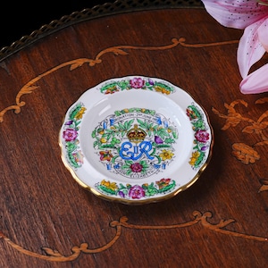 Puede incluir: Un plato conmemorativo de cer&aacute;mica blanca con un borde dorado festoneado. El centro presenta el monograma real azul EIIR, una corona y el texto Coronation 2nd June 1953 Queen Elizabeth II, rodeado de coloridas ilustraciones florales de rosas y cardos.