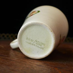 Puede incluir: Taza de cer&aacute;mica color crema con asa, con dise&ntilde;os florales en tonos rojos, naranjas y verdes. La base de la taza muestra el texto "BCW/KELSON WARE MADE IN ENGLAND" en color verde descolorido. La taza est&aacute; sobre una superficie de madera.