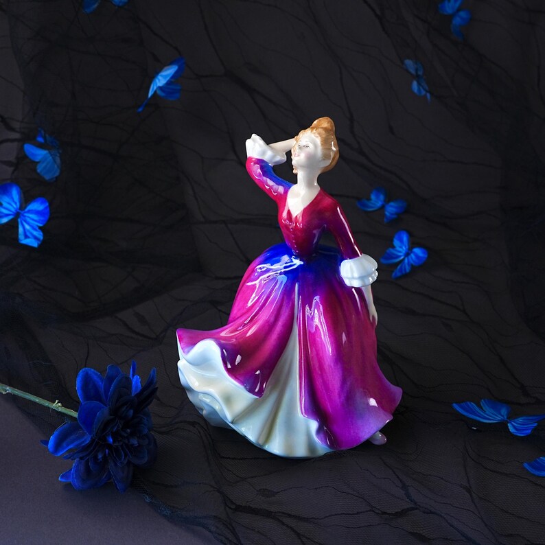 May include: A porcelain figurine of a person in a long, flowing dress that transitions from a deep magenta at the bodice to a vibrant blue and white at the hem. The figure has light skin, brown hair, and wears white ruffled cuffs on the sleeves.