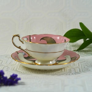 May include: A vintage teacup and saucer set. The teacup has a pink interior and a white exterior with gold trim. The saucer is pink with a matching design. The set is likely made of porcelain and is suitable for serving tea.