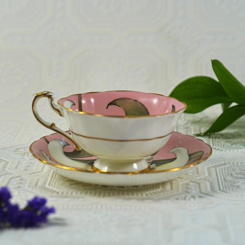 May include: A vintage teacup and saucer set. The teacup has a pink interior and a white exterior with gold trim. The saucer is pink with a matching design. The set is likely made of porcelain and is suitable for serving tea.