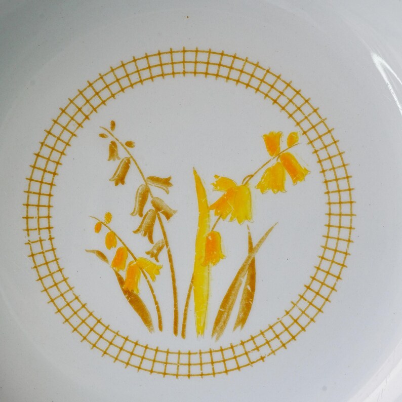 May include: White ceramic plate with a yellow and brown floral design. The flowers are inside a yellow grid circle. The plate has a simple, clean design, with a focus on the floral pattern.
