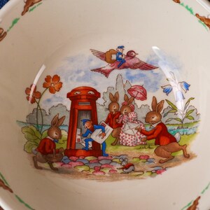May include: A cream-colored ceramic bowl featuring a colorful illustration of anthropomorphic rabbits in a garden setting. The scene depicts rabbits near a red pillar box, a bird carrying a figure, and floral accents along the bowl's rim.