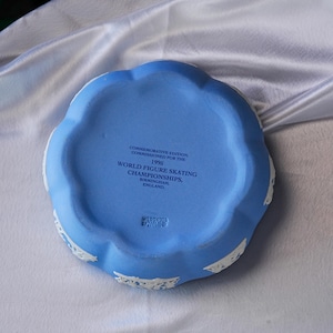 Może przedstawiać: Niebiesko-biała pamiątkowa miseczka. Miseczka Wedgwood z 1995 roku, zam&oacute;wiona na Mistrzostwa Świata w Łyżwiarstwie Figurowym w Birmingham, Anglia. Ma falisty brzeg z białymi detalami.