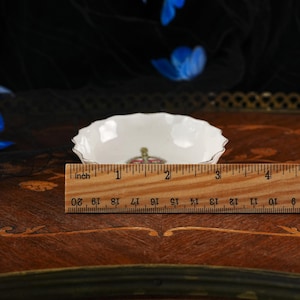 Op de afbeelding: Een kleine witte porseleinen schaal met een geschulpte rand en een kroonembleem in het midden. Een houten liniaal ligt voor de schaal en toont een diameter van ongeveer 9 cm. De schaal is voorzien van een dunne zilveren rand.