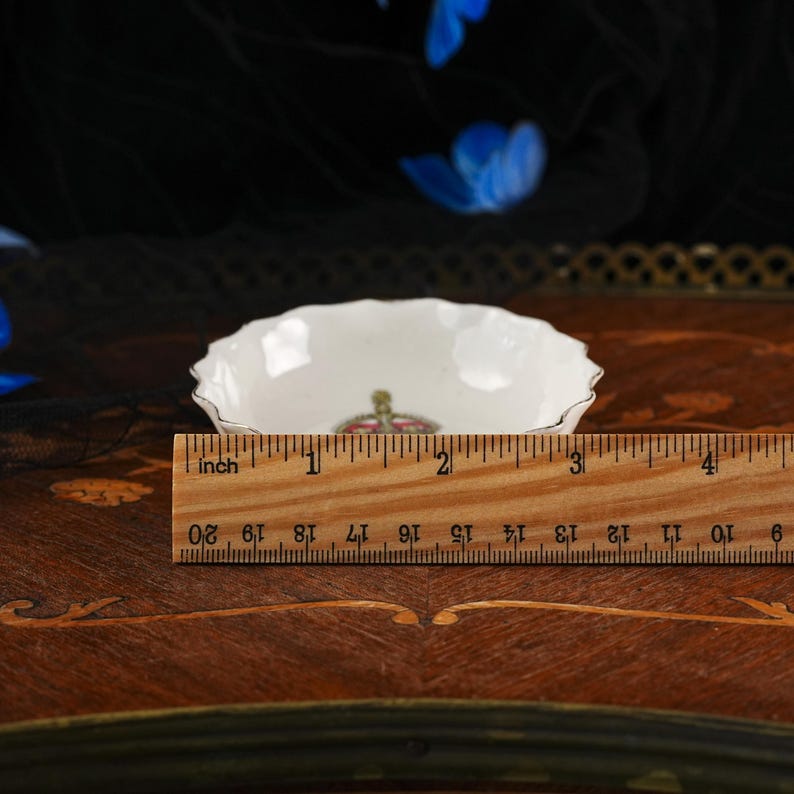 Op de afbeelding: Een kleine witte porseleinen schaal met een geschulpte rand en een kroonembleem in het midden. Een houten liniaal ligt voor de schaal en toont een diameter van ongeveer 9 cm. De schaal is voorzien van een dunne zilveren rand.