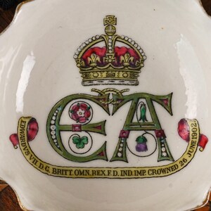 Op de afbeelding: Een witte keramische herdenkingsschaal met een gouden kroon boven de letters EA. Het ontwerp bevat een roos, klaverblad en distel, met een geel lint met de inscriptie EDVARDVS VII D.G. BRITT. OMN. REX F.D. IND. IMP. CROWNED 26 JUNE 1902.