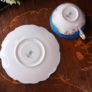 Op de afbeelding: Een vintage theekop en schotel set. De theekop is wit met een blauwe band en gouden accenten. De schotel is wit met een geschulpte rand. Beide stukken liggen op een houten oppervlak.