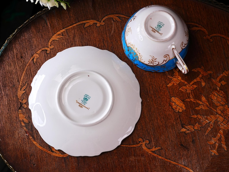 Op de afbeelding: Een vintage theekop en schotel set. De theekop is wit met een blauwe band en gouden accenten. De schotel is wit met een geschulpte rand. Beide stukken liggen op een houten oppervlak.
