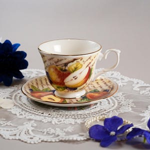 Könnte beinhalten: Weißes Tee-Set mit Tasse und Untertasse mit goldenem Rand. Die Tasse und Untertasse sind mit einem Fruchtdesign mit Birnen und Pfirsichen verziert. Der Hintergrund hat handschriftlichen Text.