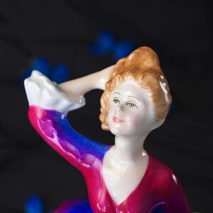 May include: A porcelain figurine of a person with light brown hair styled in an updo, wearing a vibrant dress with a deep red bodice and blue sleeves featuring white ruffled cuffs. The figure has a pale complexion, painted facial features, and a hand raised to the head, captured from the chest up in a detailed ceramic finish.