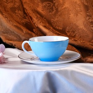 Pode incluir: Uma ch&aacute;vena e pires azul claro. A ch&aacute;vena tem interior branco e uma borda dourada. O pires &eacute; branco com um delicado padr&atilde;o floral e uma borda dourada. O conjunto est&aacute; sobre uma toalha de mesa branca.
