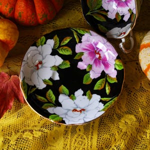 May include: A black saucer with a gold rim, adorned with white and pink floral designs and green leaves. The saucer is part of a tea set, with a matching teacup visible in the background. The set is placed on a yellow lace tablecloth.