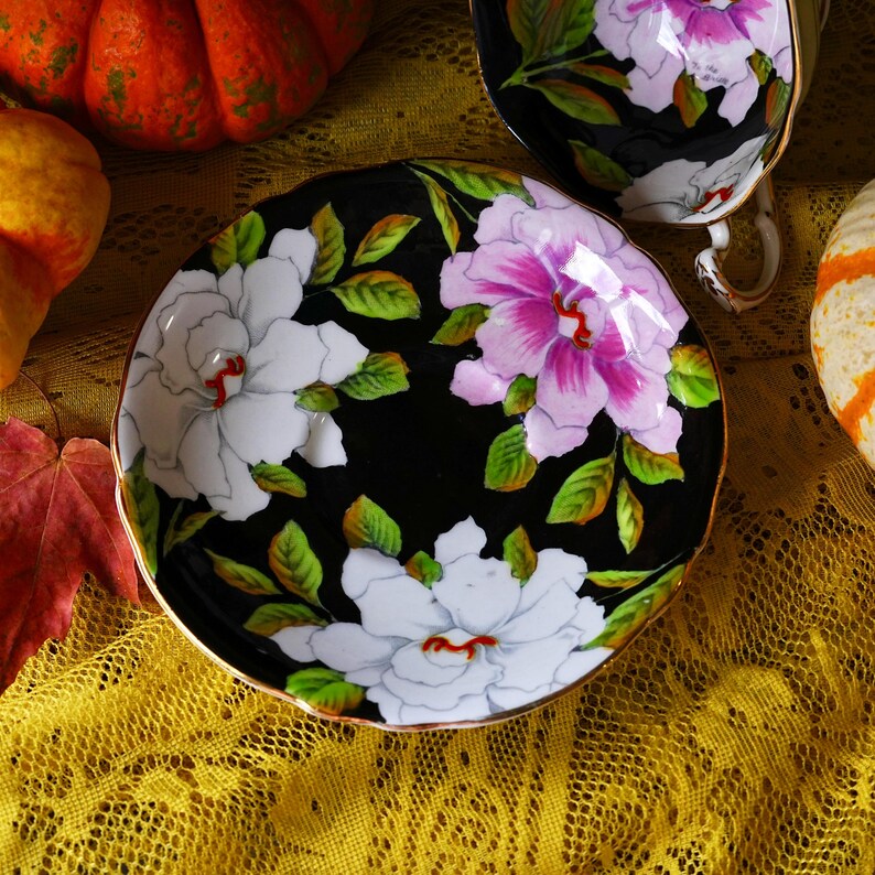 May include: A black saucer with a gold rim, adorned with white and pink floral designs and green leaves. The saucer is part of a tea set, with a matching teacup visible in the background. The set is placed on a yellow lace tablecloth.