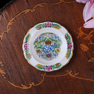 Puede incluir: Un plato conmemorativo de cer&aacute;mica blanca con un borde dorado festoneado. El centro presenta una ilustraci&oacute;n colorida de una corona, el monograma real EIIR y el texto CORONATION 2ND JUNE 1953 QUEEN ELIZABETH II, rodeado de motivos florales.