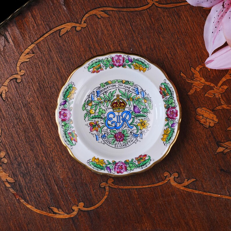 Puede incluir: Un plato conmemorativo de cer&aacute;mica blanca con un borde dorado festoneado. El centro presenta una ilustraci&oacute;n colorida de una corona, el monograma real EIIR y el texto CORONATION 2ND JUNE 1953 QUEEN ELIZABETH II, rodeado de motivos florales.