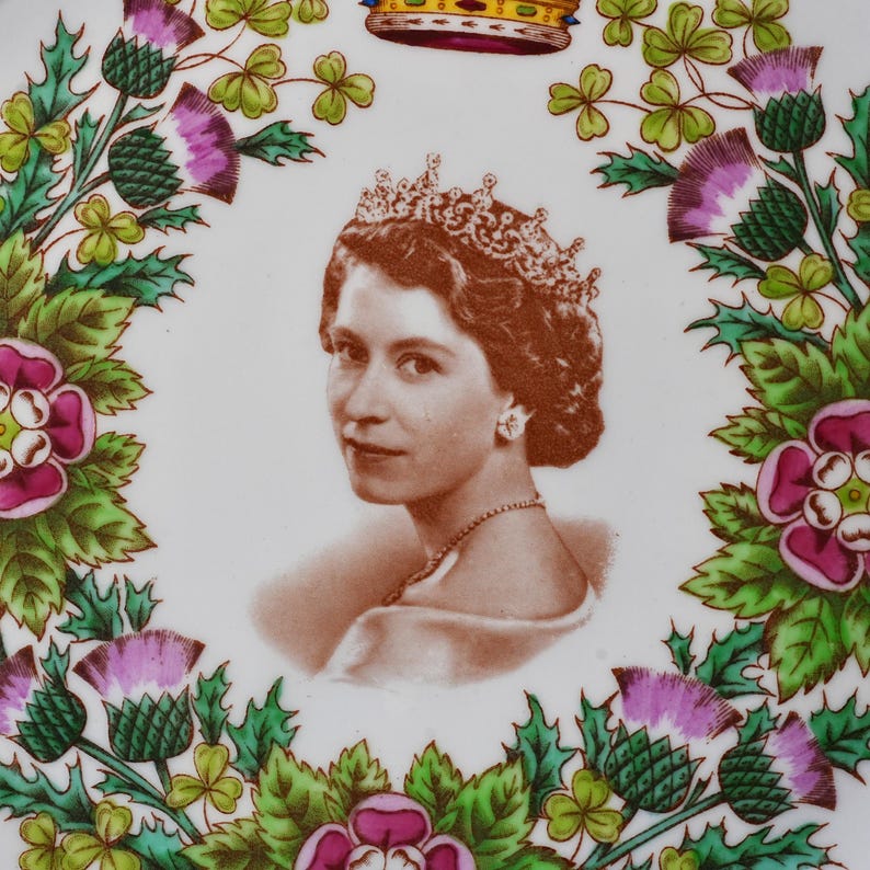 Op de afbeelding: Een wit keramieken bord met een sepia-kleurig portret van koningin Elizabeth II met een kroon en ketting. Het portret is omlijst door een kleurrijke bloemenkrans van paarse distels, roze rozen en groene klaverbladeren met een kroon.