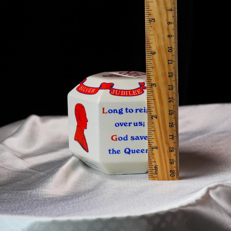 Op de afbeelding: Witte keramische herdenkingsdoos met een rood en blauw ontwerp. De doos heeft de tekst "SILVER JUBILEE" en "Long to reign over us; God save the Queen." De doos is ongeveer 7,6 cm hoog.