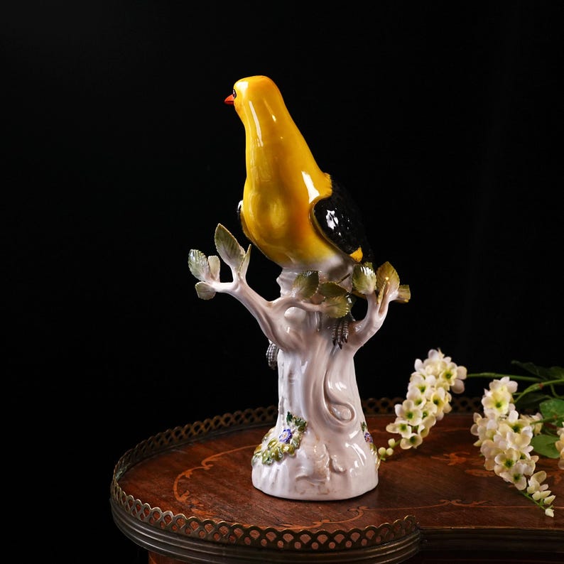 May include: A yellow and black ceramic bird figurine perched on a white tree branch with green leaves. The bird has a red beak and is set on a decorative base with floral accents. The figurine is displayed on a wooden table.