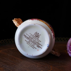 Puede incluir: Una crema de porcelana blanca con un asa y borde decorativos. La crema tiene un dise&ntilde;o floral con detalles dorados. La parte inferior de la crema tiene el texto "King George VI & Queen Elizabeth" y "Made in England."