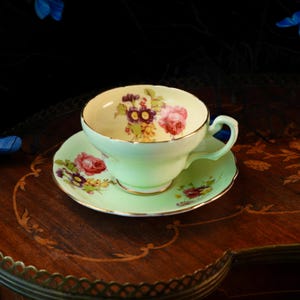 Puede incluir: Un juego de taza de té y platillo de porcelana en color verde menta pálido con un delicado patrón floral de rosas rosas y pequeñas flores moradas y amarillas. El borde de la taza y del platillo está acabado con un fino ribete dorado.