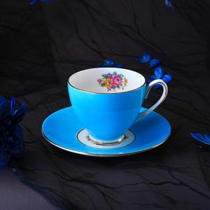 Op de afbeelding: Een fel turquoise theekop en schotel set met een goudkleurige rand en handvat. De binnenkant van de kop en het midden van de schotel zijn versierd met een klein, kleurrijk boeket van roze, gele en paarse bloemen.