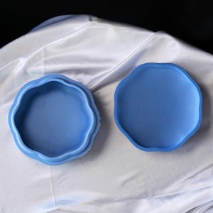 Może przedstawiać: Dwuczęściowa, niebieska ceramiczna miseczka na drobiazgi o falistym, organicznym kształcie. Miseczka jest otwarta, odsłaniając wnętrze, a pokrywka jest umieszczona obok. Tło stanowi biała, jedwabista tkanina.
