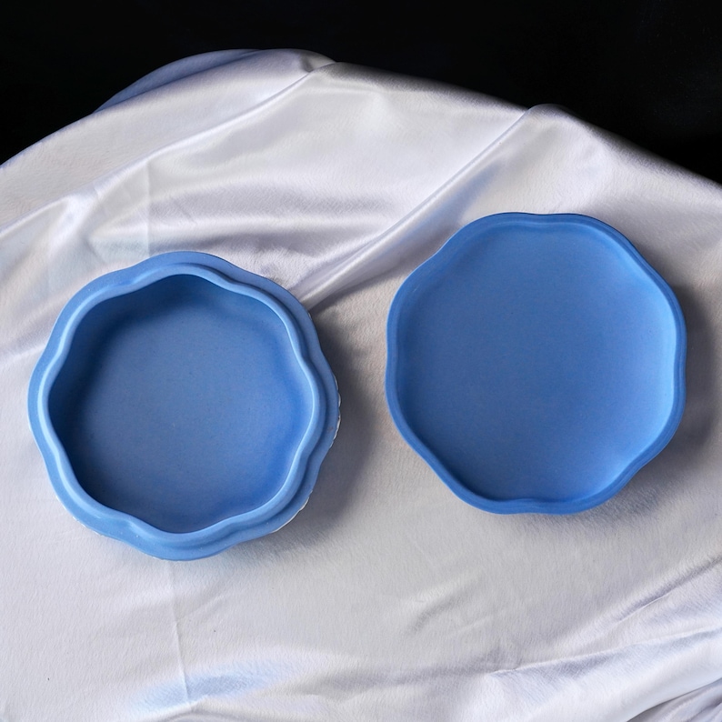 Może przedstawiać: Dwuczęściowa, niebieska ceramiczna miseczka na drobiazgi o falistym, organicznym kształcie. Miseczka jest otwarta, odsłaniając wnętrze, a pokrywka jest umieszczona obok. Tło stanowi biała, jedwabista tkanina.