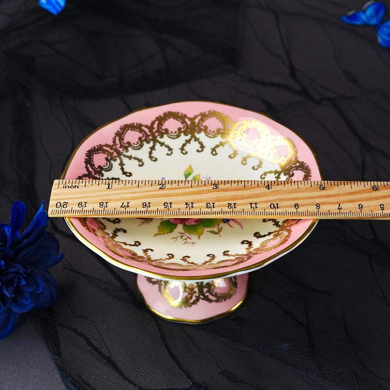 Puede incluir: Un cuenco de pedestal de porcelana rosa y crema con un motivo floral central y un borde de filigrana dorada. Una regla de madera colocada sobre el cuenco muestra un di&aacute;metro de aproximadamente 14 cm. El cuenco tiene un borde festoneado.