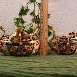 May include: A decorative tea set with a teapot and creamer. The teapot is approximately 6 inches tall, and the creamer is about 3 inches tall. Both pieces feature a floral design with gold trim and accents. The set is on a green surface.