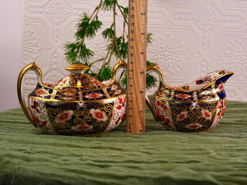 May include: A decorative tea set with a teapot and creamer. The teapot is approximately 6 inches tall, and the creamer is about 3 inches tall. Both pieces feature a floral design with gold trim and accents. The set is on a green surface.
