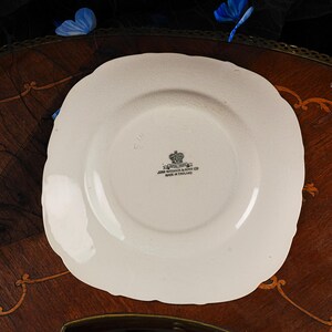 May include: An off-white ceramic saucer with a scalloped edge. The centre features a black stamp with a crown icon and the text: ROYAL IVORY, JOHN MADDOCK & SONS LTD, MADE IN ENGLAND. The saucer has a smooth, glossy finish and a circular indentation.