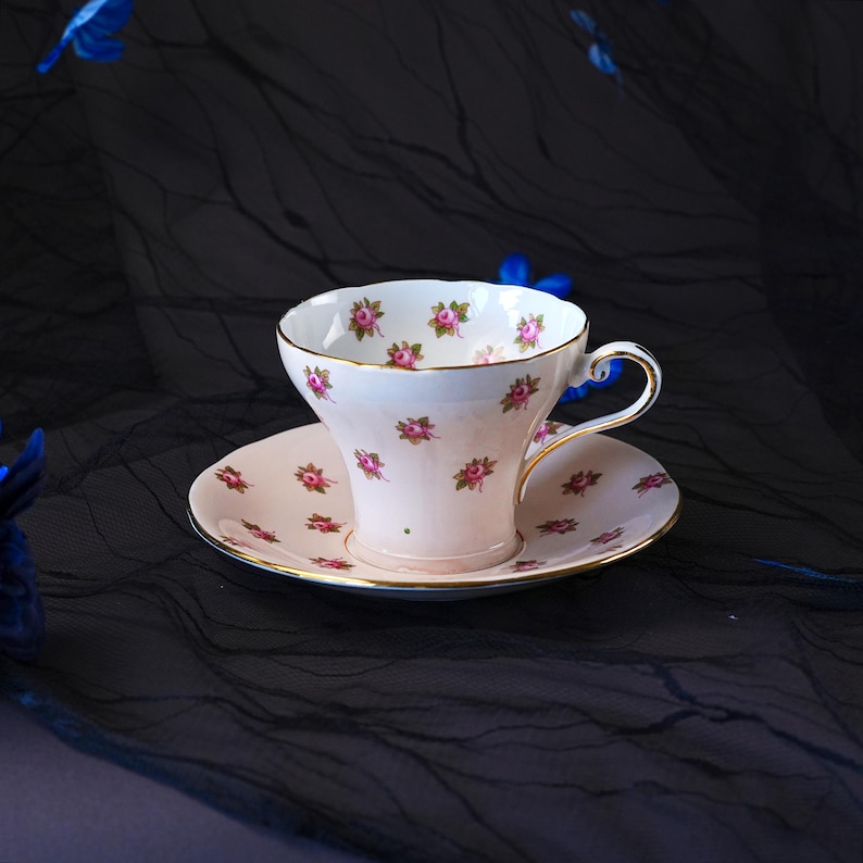 Może przedstawiać: Zestaw filiżanki ze spodkiem z porcelany o jasnokremowej barwie, ozdobiony powtarzającym się wzorem małych r&oacute;żowych pąk&oacute;w r&oacute;ż i zielonych liści. Krawędzie filiżanki oraz spodka wykończone są cienką złotą obw&oacute;dką, filiżanka posiada wygięte ucho.