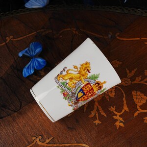 Puede incluir: Una taza de cer&aacute;mica blanca con un le&oacute;n dorado y un escudo real colorido con el texto Coronation AD 1911. La taza tiene un borde dorado fino en la parte superior y una forma cil&iacute;ndrica c&oacute;nica, mostrando un dise&ntilde;o her&aacute;ldico detallado en rojo, azul y oro.