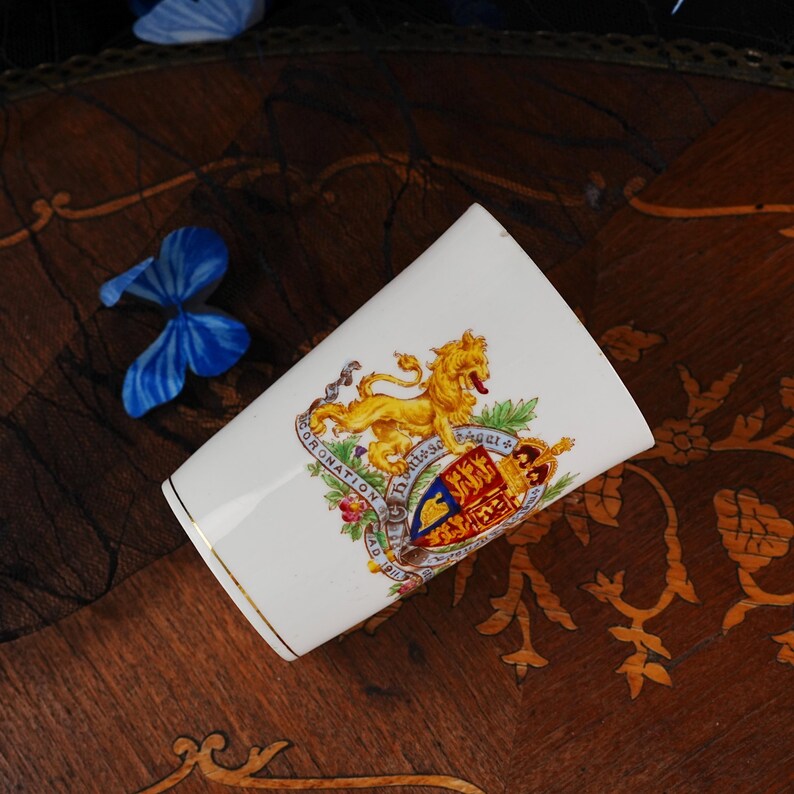Puede incluir: Una taza de cer&aacute;mica blanca con un le&oacute;n dorado y un escudo real colorido con el texto Coronation AD 1911. La taza tiene un borde dorado fino en la parte superior y una forma cil&iacute;ndrica c&oacute;nica, mostrando un dise&ntilde;o her&aacute;ldico detallado en rojo, azul y oro.