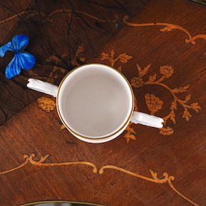May include: A white porcelain cup with two handles and a gold-trimmed rim, viewed from directly above. The cup features a smooth, plain interior and exterior surface, with the gold band encircling the top edge of the circular vessel.