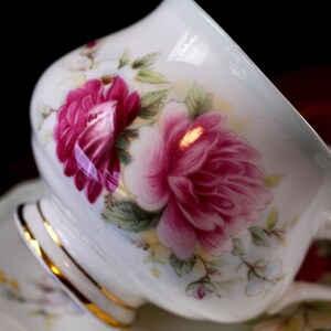 May include: Close-up of a white teacup and saucer set with a floral design. The teacup features a large, vibrant pink peony with green leaves. The rim of the cup and saucer are trimmed with gold.