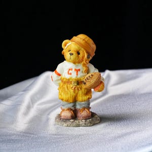 Op de afbeelding: Een gedetailleerd beeldje van een teddybeer in een football-uniform. De beer draagt een helm en houdt een football vast. Het uniform is wit met oranje accenten en de letters "CT". De beer staat op een kleine, getextureerde basis.