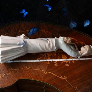 K&ouml;nnte beinhalten: Eine Porzellanfigur einer Person mit Hut und langem Mantel, die horizontal neben einem wei&szlig;en Ma&szlig;band liegt, das eine L&auml;nge von etwa 35,5 cm anzeigt. Die Figur hat eine gl&auml;nzende Oberfl&auml;che in Wei&szlig; und Hellblau.