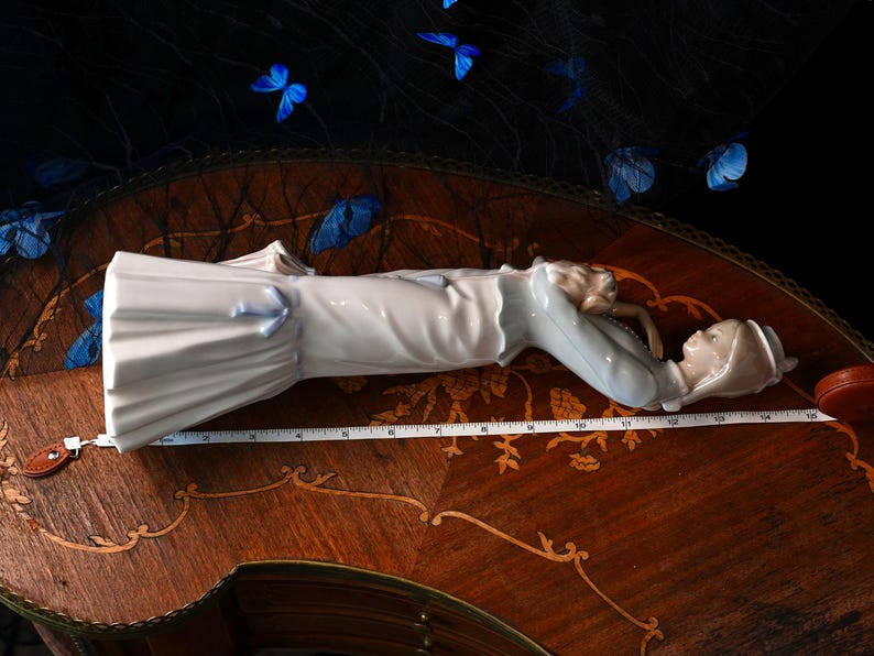 K&ouml;nnte beinhalten: Eine Porzellanfigur einer Person mit Hut und langem Mantel, die horizontal neben einem wei&szlig;en Ma&szlig;band liegt, das eine L&auml;nge von etwa 35,5 cm anzeigt. Die Figur hat eine gl&auml;nzende Oberfl&auml;che in Wei&szlig; und Hellblau.