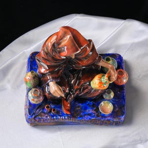 May include: A ceramic figurine of a figure in a brown robe, surrounded by small, colourful vases. The figure sits on a blue base with a textured surface. The vases are in various colours, including green, orange, and yellow.