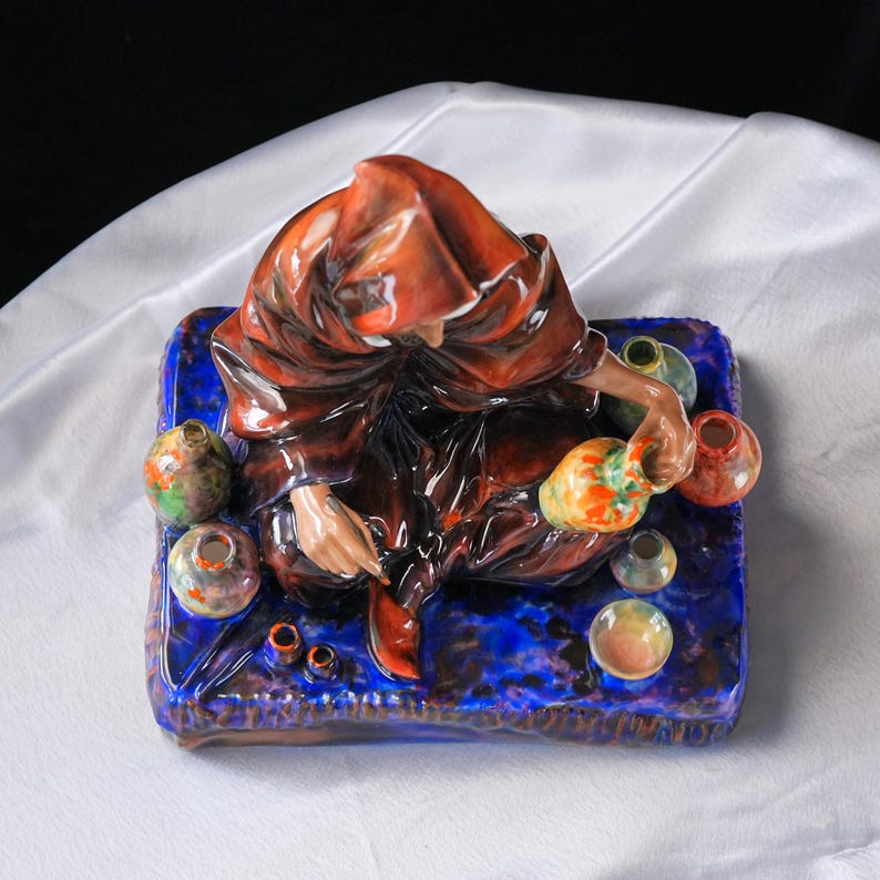 May include: A ceramic figurine of a figure in a brown robe, surrounded by small, colourful vases. The figure sits on a blue base with a textured surface. The vases are in various colours, including green, orange, and yellow.