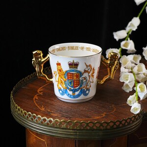 Puede incluir: Una taza de porcelana blanca con dos asas, borde dorado y un escudo real colorido con un le&oacute;n y un unicornio. El borde interior muestra el texto THE GOLDEN JUBILEE OF HER MAJESTY. Las asas tienen forma de figuras de color dorado.