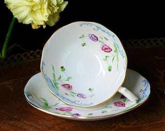 May include: White teacup and saucer set with a floral design in pink, purple, and green. The cup and saucer have a gold trim and a light blue accent. The set is on a wooden surface.