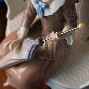 K&ouml;nnte beinhalten: Eine Porzellanfigur, die eine Person in einem braunen lang&auml;rmeligen Oberteil mit wei&szlig;em Kragen und hellblauer Schleife zeigt. Die Figur h&auml;lt einen geschlossenen hellrosa Regenschirm mit blauem Griff und einem d&uuml;nnen Goldstab, der mit einer cremefarbenen Perle verziert ist.
